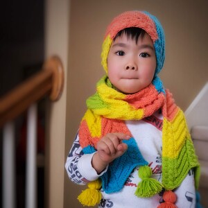 May include: A young child wearing a colorful crocheted hat and scarf. The hat and scarf are made with a multi-colored yarn in shades of pink, yellow, blue, and green. The child is wearing a white shirt with a graphic print and dark pants.
