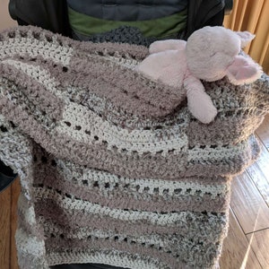 May include: A handmade crochet blanket in shades of white, gray, and taupe, draped over a chair. A pink plush bunny rests on the blanket. The blanket has a textured, open weave design.