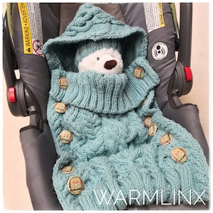 May include: A teal blue knitted baby swaddle with a hood and owl-shaped buttons. The swaddle is in a car seat and has the text "WARMLINX" on the bottom.