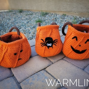 May include: Three orange crochet pumpkin-shaped treat bags with brown handles. One bag has a sad face, another has a spider, and the third has a smiling jack-o'-lantern face. The bags are sitting on a brick surface.