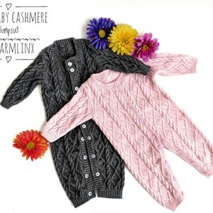 May include: Two knitted baby onesies, one gray and one pink, with button closures. The onesies have a cable knit pattern. The onesies are laid out on a white surface with flowers.