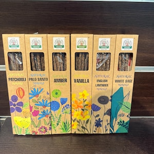 May include: Six boxes of natural incense sticks with floral designs and the names Patchouli, Palo Santo, Amber, Vanilla, English Lavender, and White Sage. The boxes are brown with a clear window showing the incense.