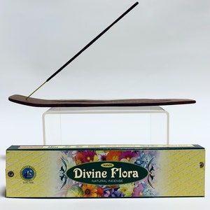 May include: A dark brown wooden incense stick holder with a single incense stick inserted, positioned above a rectangular box of Nandi Divine Flora natural incense featuring a colorful floral pattern and the text Divine Flora Natural Incense.