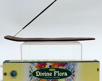 May include: A dark brown wooden incense stick holder with a single incense stick inserted, positioned above a rectangular box of Nandi Divine Flora natural incense featuring a colorful floral pattern and the text Divine Flora Natural Incense.