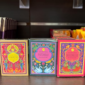 May include: Three rectangular boxes of Golden India high perfume cones. The left box is gold with red accents and Frankincense text. The middle box is dark green with Patchouli text. The right box is pink with Dragonblood text and floral art.