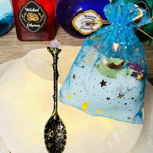 May include: An antique bronze-colored spoon with a floral design on the bowl and a crystal at the top. A blue drawstring bag with gold stars and moons is next to the spoon. Bottles with labels that read "Wicked Thorns" and "Hocus Focus" are in the background.