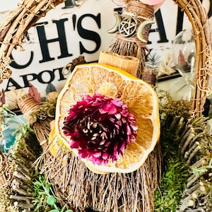 May include: A small broom with a dried orange slice and a flower on top, decorated with a pentagram charm and a moon symbol. The broom is in a woven wreath with moss and twigs.