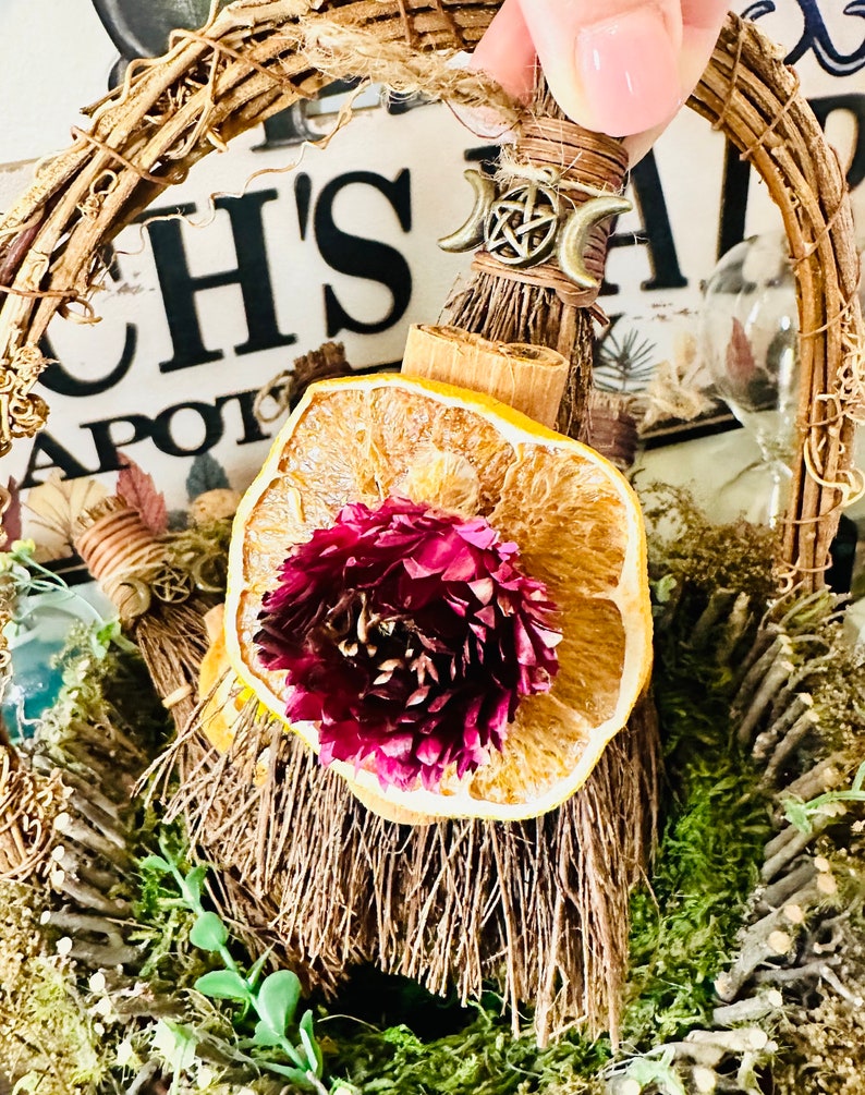 May include: A small broom with a dried orange slice and a flower on top, decorated with a pentagram charm and a moon symbol. The broom is in a woven wreath with moss and twigs.