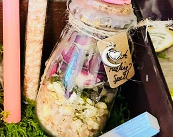 May include: A glass jar filled with dried flowers, crystals, and herbs, sealed with a cork and pink wax. A small tag with a heart and the words "making spell" hangs from the jar. A pink candle and a clear tube with contents are visible.