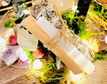 Può includere: Un fascio di salvia bianca, una bacchetta di selenite e un bastoncino di palo santo legati con spago. Gli oggetti sono circondati da cristalli, muschio e fiori secchi. Le luci delle fate aggiungono un bagliore caldo alla composizione.