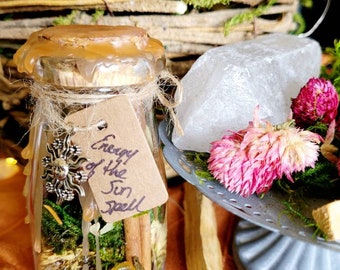 May include: A small glass bottle with a cork stopper and a brown paper tag that reads "Energy of the Sun Spell". The bottle is filled with various herbs, crystals, and other natural ingredients. The bottle is sitting on a wooden surface.