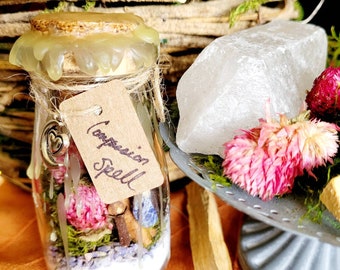 May include: A small glass jar with a cork stopper and a handwritten tag that reads "Compassion Spell". The jar is filled with dried flowers, herbs, and crystals. The jar is sitting on a wooden surface with a piece of selenite and a palo santo stick.