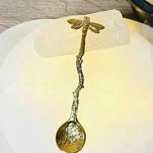 Op de afbeelding: Een gouden lepel met een bij-ontwerp op de steel. De steel is gevormd als een takje. De lepel ligt op een wit oppervlak.