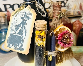 May include: A black glass bottle with a cork stopper and a tag featuring a witch stirring a cauldron. The bottle is surrounded by a small broom, two black candles, a glass vial filled with dried herbs and black sand, and a gold spoon with a crystal tip.