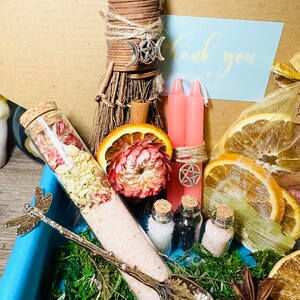 May include: A witchy gift box with a small broom, pink candles, dried orange slices, a glass bottle with pink bath salts, and a silver spoon with a dragonfly handle. The box also includes a thank you card and a small bag of dried herbs.