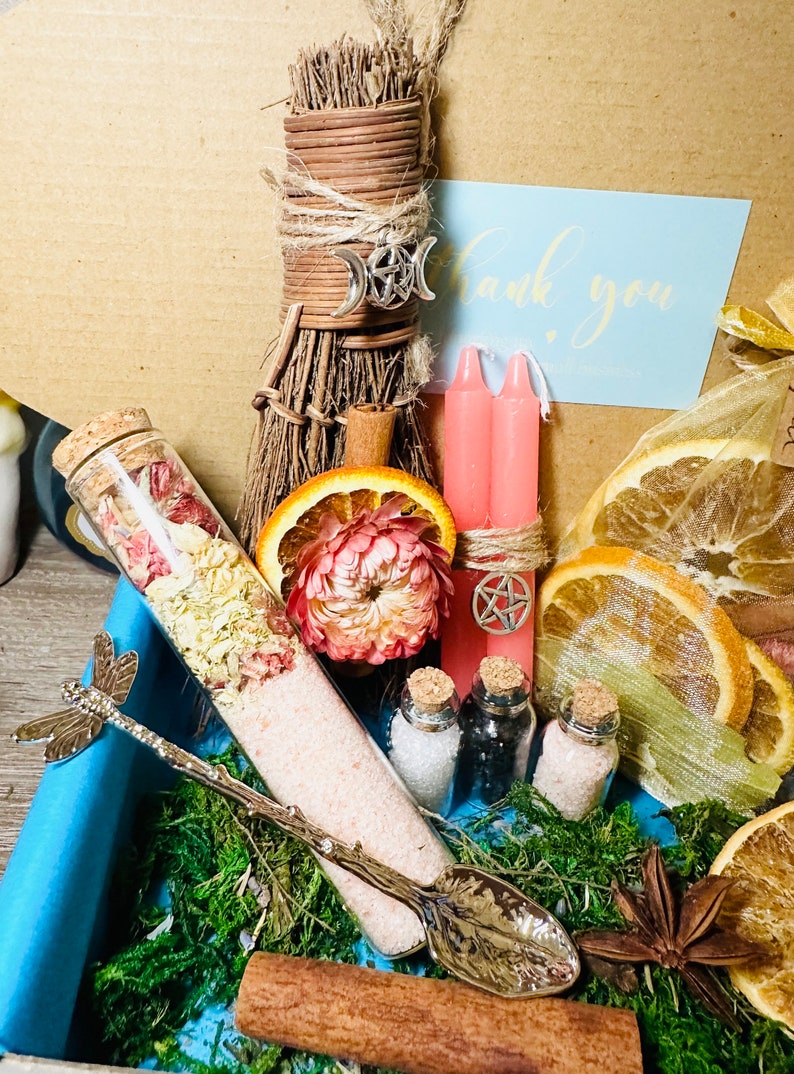 May include: A witchy gift box with a small broom, pink candles, dried orange slices, a glass bottle with pink bath salts, and a silver spoon with a dragonfly handle. The box also includes a thank you card and a small bag of dried herbs.