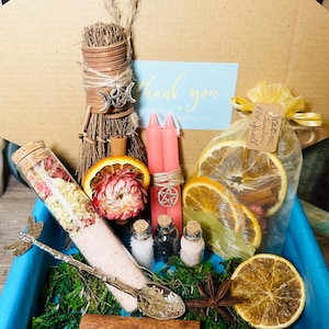 May include: A gift box filled with a variety of witchy items, including a small broom, pink candles, a bag of dried orange slices and spices, a glass tube filled with bath salts, and a small spoon. The box is lined with moss and has a blue lid.