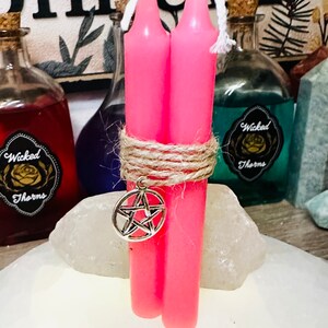 May include: Two pink candles tied together with twine and a silver pentagram charm. The candles are on a white surface.