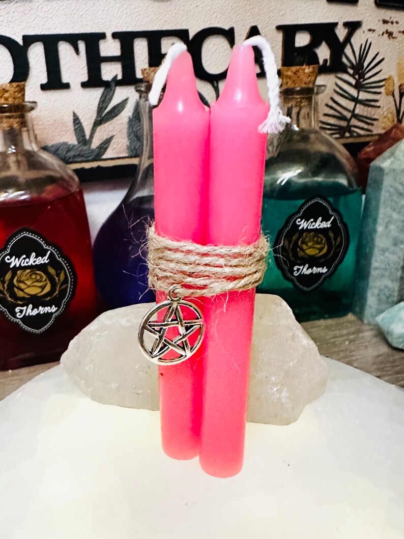May include: Two pink candles tied together with twine and a silver pentagram charm. The candles are on a white surface.
