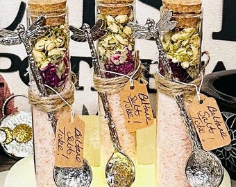 Puede incluir: Tres botellas de vidrio llenas de sal rosa y flores secas, cada una con un tapón de corcho y una cuchara de libélula. Las botellas están atadas con hilo y tienen etiquetas que dicen "Beltane Salt Tobe!".