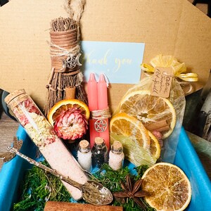 May include: A witchy gift box with a variety of items, including a small broom, pink candles, dried orange slices, a small bottle of bath salts, a spoon, and a small bag of herbs. The box is lined with moss and has a blue liner.