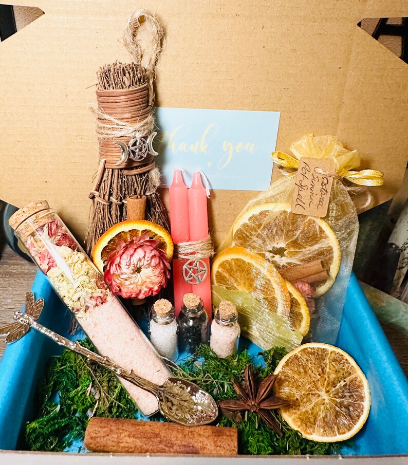 May include: A witchy gift box with a variety of items, including a small broom, pink candles, dried orange slices, a small bottle of bath salts, a spoon, and a small bag of herbs. The box is lined with moss and has a blue liner.