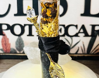 May include: A clear glass bottle filled with dried yellow flowers and black granules, topped with a cork. A gold-colored leaf-shaped charm and a crystal-topped wand are attached with a black ribbon. The bottle is displayed on a white surface.