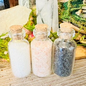May include: Three small glass bottles with cork tops filled with different types of salt. The bottles are arranged in a row from left to right: white salt, pink Himalayan salt, and black salt.