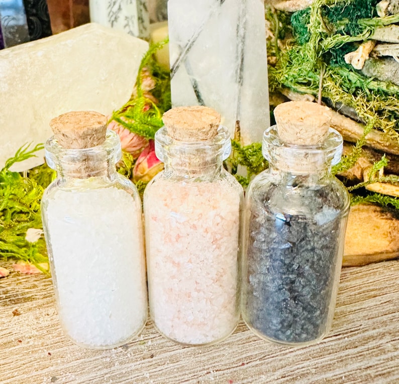 May include: Three small glass bottles with cork tops filled with different types of salt. The bottles are arranged in a row from left to right: white salt, pink Himalayan salt, and black salt.