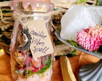 May include: A small glass jar with a pink wax seal and a brown paper tag that reads "Positive Vibes Spell". The jar is filled with pink Himalayan salt, dried herbs, and other natural ingredients.