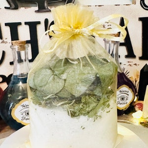 Puede incluir: Una bolsa blanca con un cordón de ajuste amarillo y una cinta amarilla atada alrededor. La bolsa está llena de hojas verdes y cristales blancos. La bolsa está sentada sobre una superficie blanca. El texto "Hocus Focus" es visible en una pequeña etiqueta adjunta a la bolsa.