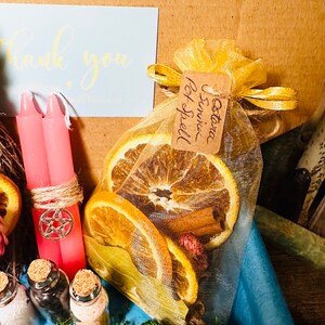 May include: A yellow organza bag filled with dried orange slices, cinnamon sticks, and pink flower buds. The bag is tied with a yellow ribbon and has a brown tag that reads "Autumn & Spice Spell".