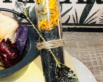 May include: A clear glass vial filled with dried yellow flowers and dark granules, sealed with a cork stopper. A decorative spoon with a floral design is attached with twine. The vial is part of a collection of natural elements.