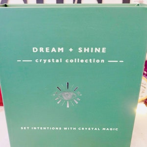 Puede incluir: Una caja verde con el texto "DREAM + SHINE - crystal collection -" y un símbolo de ojo plateado. El texto "SET INTENTIONS WITH CRYSTAL MAGIC" está en la parte inferior de la caja.