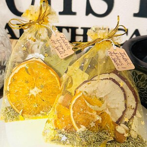 May include: Two small, gold mesh bags tied with gold ribbon, each containing dried orange slices, herbs, and a tag that reads "Imbolc Simmer Pot Spell!". The bags are decorated with gold stars and sit on a white surface.