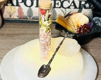 May include: A glass vial filled with pink Himalayan salt, dried flowers, and herbs. The vial is on a white selenite plate with a silver dragonfly spoon.