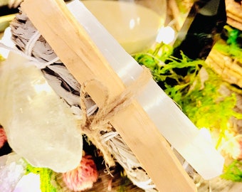 Può includere: Un fascio di bastoncini di salvia bianca per la purificazione, legati con spago e una bacchetta di selenite. Il fascio poggia su un letto di muschio e piccoli cristalli.