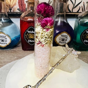 May include: A glass jar with a cork lid filled with pink bath salts, dried flowers, and a silver dragonfly spoon. The jar is labeled "Wicked Thorn" and is sitting on a white surface with other jars labeled "Focus" and "The"
