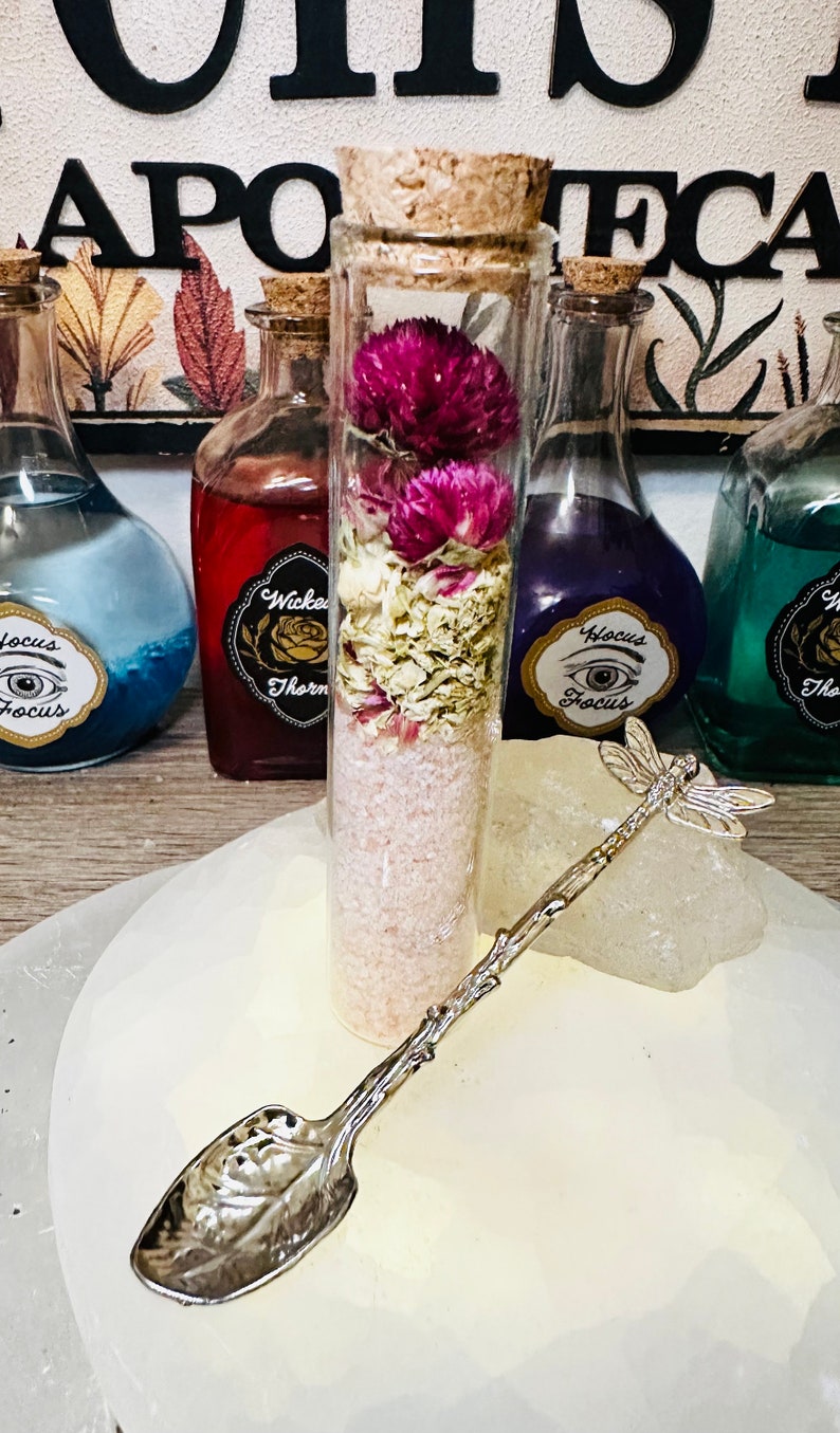 May include: A glass jar with a cork lid filled with pink bath salts, dried flowers, and a silver dragonfly spoon. The jar is labeled "Wicked Thorn" and is sitting on a white surface with other jars labeled "Focus" and "The"