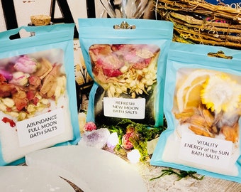 May include: Three clear plastic bags with blue borders contain bath salts. The bags are labeled with the text "Abundance Full Moon Bath Salts", "Refresh New Moon Bath Salts", and "Vitality Energy of the Sun Bath Salts".