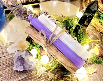 Può includere: Un set di candele viola e bianche con un bastoncino di salvia, legato con spago. Il set è circondato da muschio, cristalli e luci fatate.