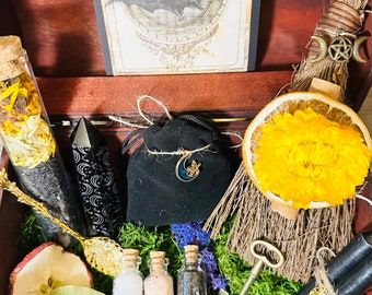 May include: A wooden box filled with various witchy items, including a dried orange slice, a small black pouch with a crescent moon charm, a glass vial with dried flowers, a black crystal, a gold spoon, three small glass vials with white, pink, and black substances, a key, and a broom with a pentagram charm.