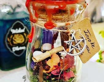 May include: A glass jar filled with dried flowers, herbs, and crystals, sealed with red wax and twine. A tag with the words "Invoke now" hangs from the twine. A silver pentacle charm is attached to the jar.
