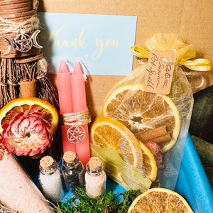 May include: A witchy gift box with a variety of items, including a small broom, two pink candles, a small bag of dried orange slices and cinnamon sticks, three small glass bottles with cork tops, a small silver spoon, a star anise, and a thank you note.