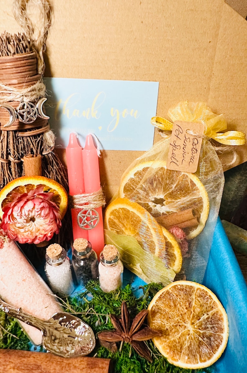May include: A witchy gift box with a variety of items, including a small broom, two pink candles, a small bag of dried orange slices and cinnamon sticks, three small glass bottles with cork tops, a small silver spoon, a star anise, and a thank you note.
