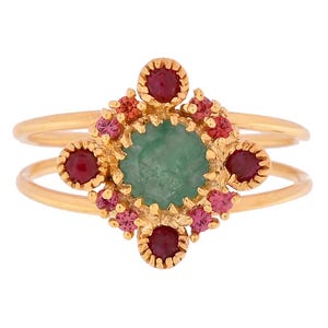 Op de afbeelding: Een goudkleurige ring met een dubbele band. De ring heeft een centrale, lichtgroene edelsteen, omringd door kleinere rode en roze edelstenen. Het ontwerp is symmetrisch en sierlijk.