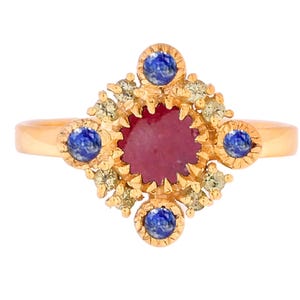 Op de afbeelding: Een gouden ring met een centrale, ronde, rode edelsteen, omringd door kleinere blauwe en heldere edelstenen. De ring heeft een gedetailleerd, sierlijk ontwerp met een band die naar achteren taps toeloopt. De ring staat tegen een witte achtergrond.