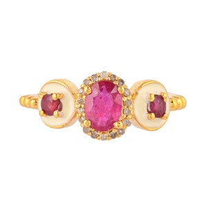 Puede incluir: Un anillo de oro con una gran piedra central ovalada de rubí rodeada de pequeños diamantes. El anillo tiene dos piedras laterales con un centro de rubí y esmalte blanco. Es una joya.