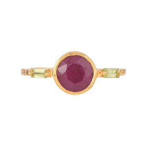 May include: A gold ring featuring a large, round, red gemstone surrounded by smaller, green gemstones and tiny white diamonds.