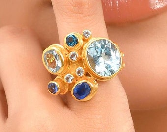 May include: A gold-toned ring featuring a cluster of various-sized gemstones. The ring showcases a large, light blue gemstone at the center, surrounded by smaller blue, white, and light blue stones. The ring is worn on a finger.
